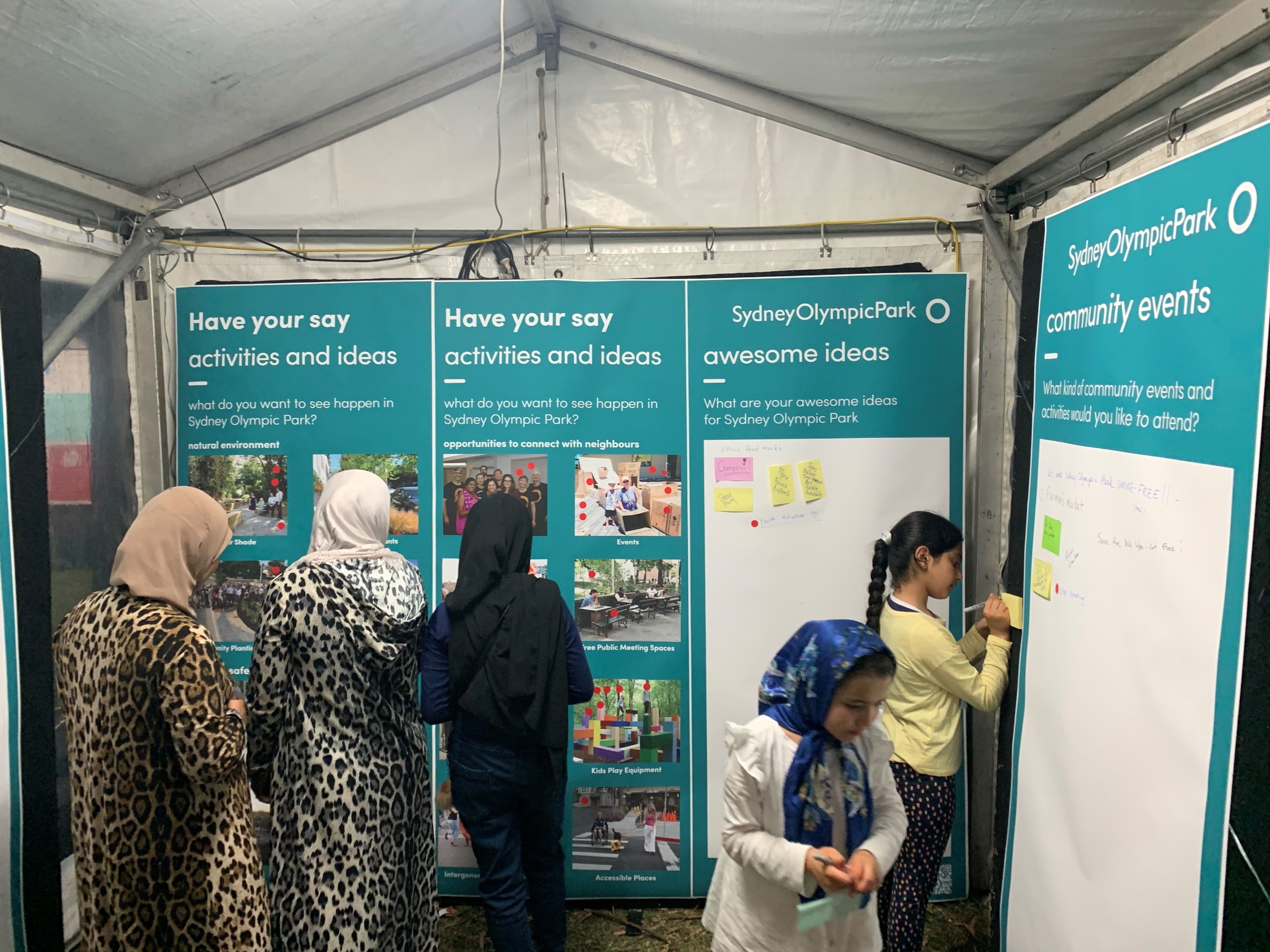 Three women looking at the activities and ideas pictures on the ideas boards in a pop-up stall. Two young girls are next to them writing ideas on post it notes to go on the other boards.