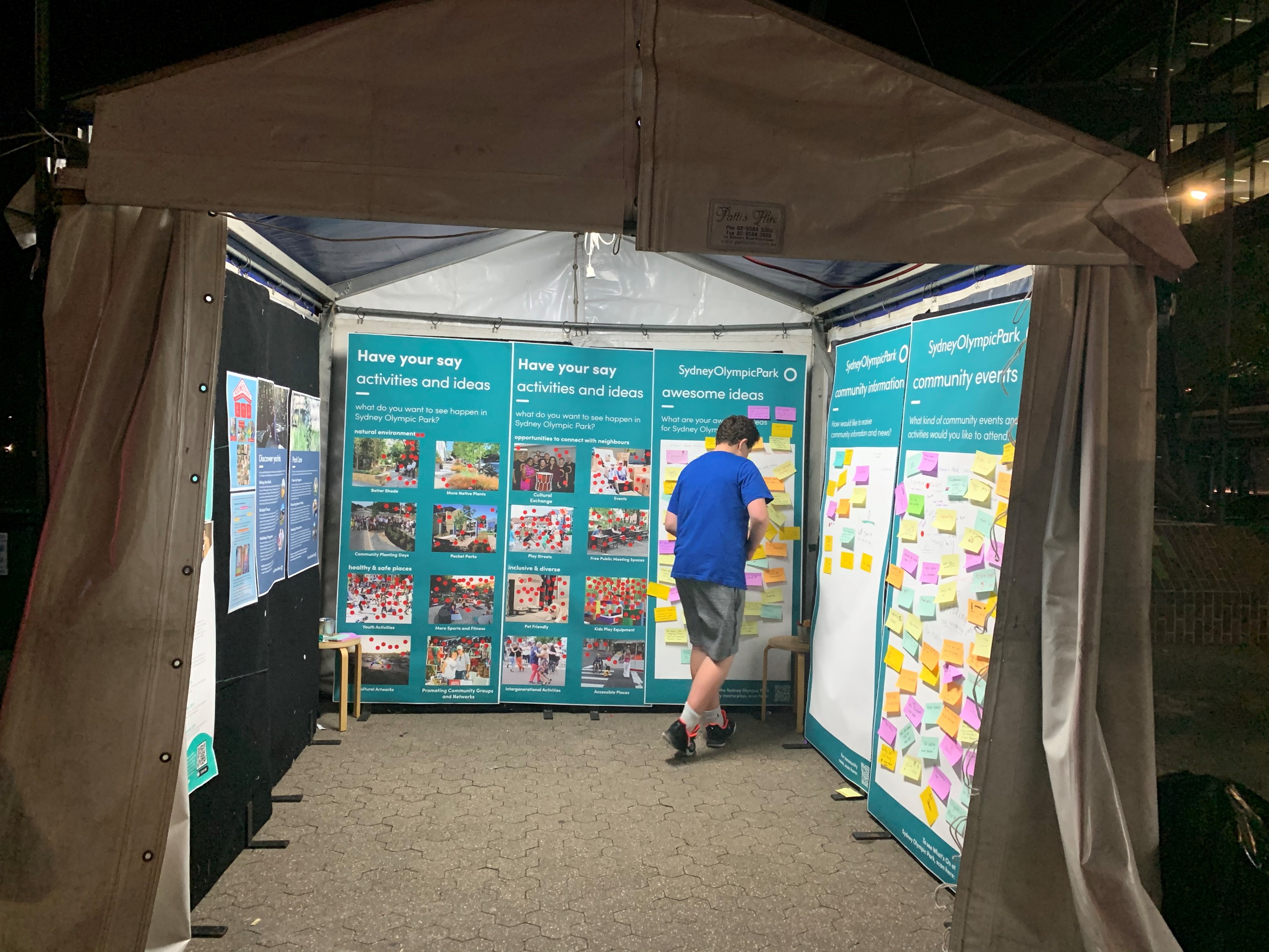 The pop-up stall viewed from outside at night, with a young man standing in front of the ideas boards with post-it notes all over them, looking like he is about to add a note.