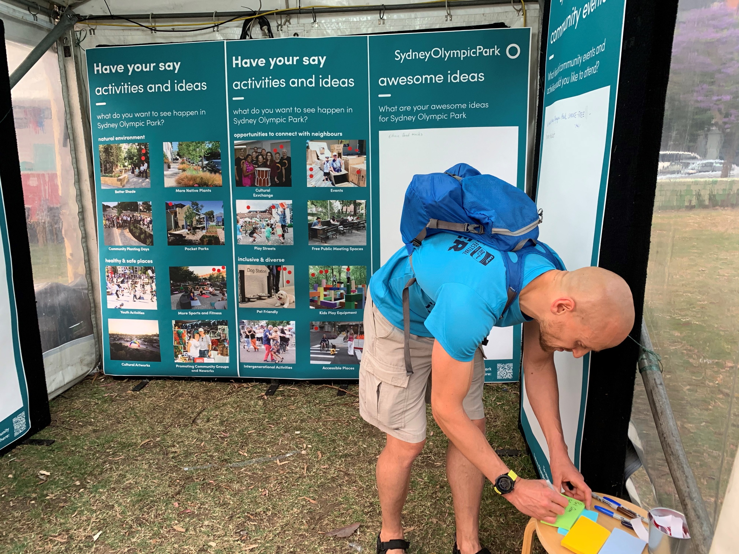 A man bending down and writing on a post-it note at the front of a stall in a pop-up tent, behind him are the ideas boards which include pictures of ideas and spaces to add the post-it notes.  